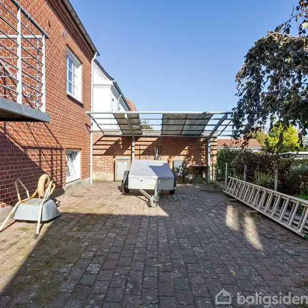 En murstensbygning har en gårdsplads med brostensbelægning. En trillebør står ved siden af en hvid trailer. Der er en stige langs en hæk, og et halvtag dækker en del af området.