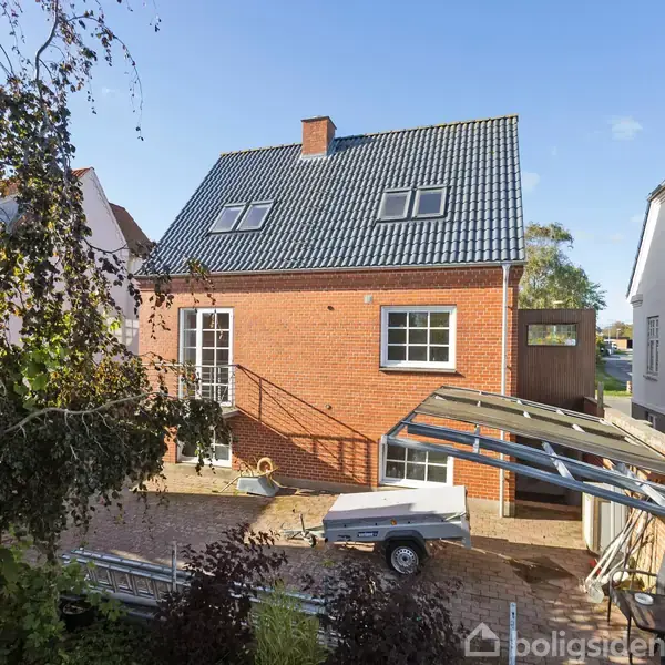 Rødt murstenhus med sort tag står stille i en solrig gårdhave med trailer og overdækket cykelskur mellem to andre huse. Træer omgiver området.