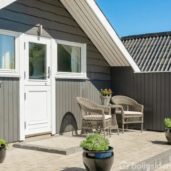 Et gråt træhus med et hvidt dørparti på en terrasse. To fletstole står ved et rundt bord. Blomsterkrukker dekorerer den flisebelagte terrasse under en klar, blå himmel.