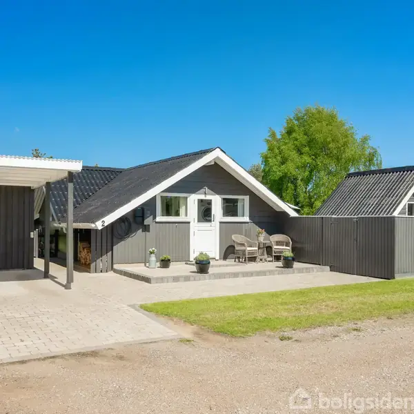 Et gråt træhus med hvide detaljer står ved siden af en carport. Foran huset er der stole og planter. Omgivet af en grøn græsplæne og træer under en blå himmel.