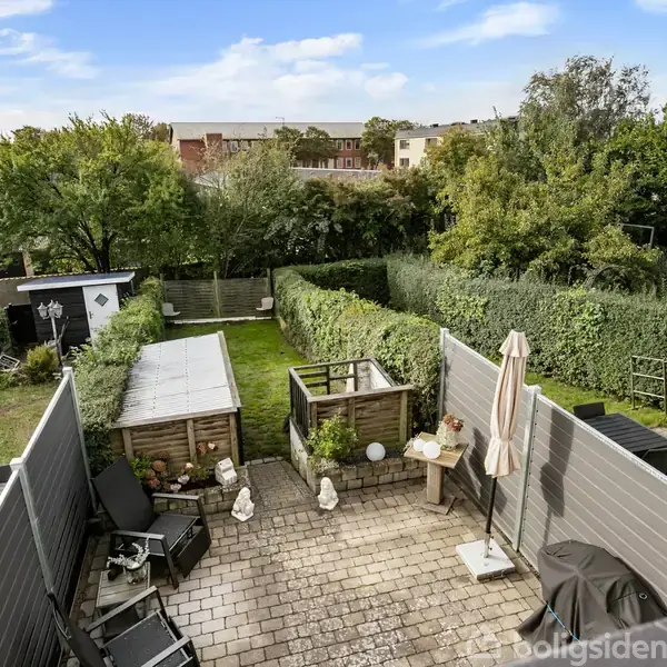 Terrasse med flisebelagt gulv, havemøbler og parasol, omkranset af lave hegn. Græsplæne med buske og træer i baggrunden, klart vejr med blå himmel og få skyer.
