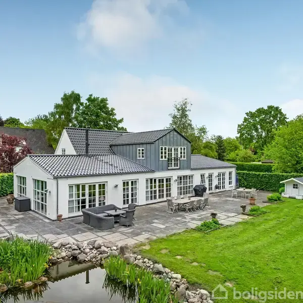 Hus med hvid facade og gråt tag står stille i en frodig have med terrasse og lille dam. Omkring er der græsplæne og træer.