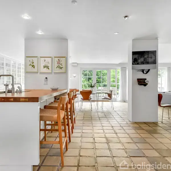 Køkken med hvidt interiør og barstole ved køkkenø, der står i et rummeligt og lyst rum med klinkegulv. I baggrunden ses et spisebord og store vinduer med udsigt til grønne træer.