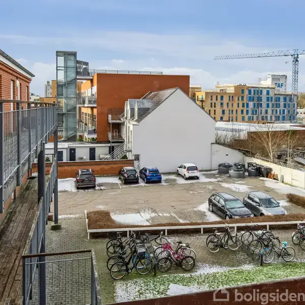 Cykler står parkeret på stativer i en indhegnet gård med få biler parkeret. Omgivet af murstensbygninger og moderne byggeri ses i baggrunden med en byggekran i det fjerne.