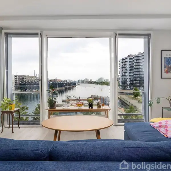 Blå sofa står i en stue med store vinduer, der giver udsigt til en havn med moderne bygninger. En træbord er placeret foran sofaen, omgivet af planter og dekorationer.