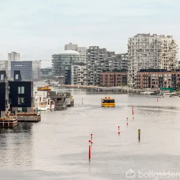 En gul færge sejler i en bred kanal, omgivet af moderne bygninger i forskellige arkitektoniske stilarter, med boligblokke og kontorer langs bredderne.
