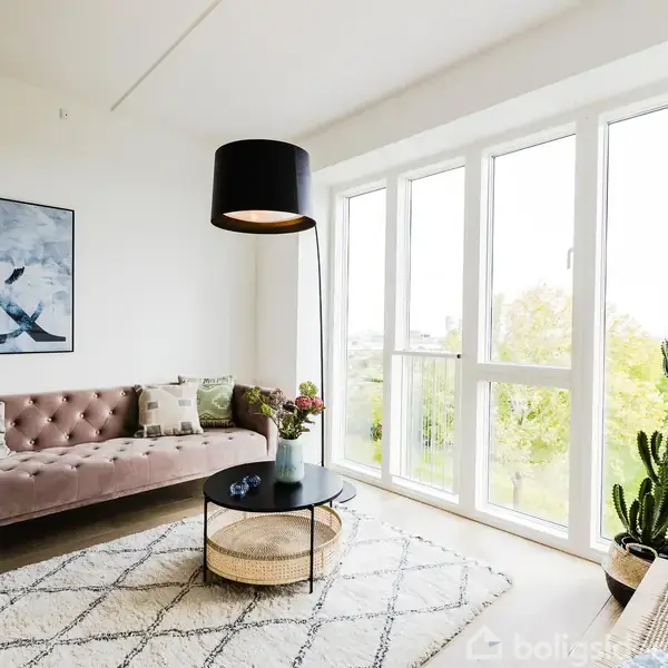 Sofa placeret i lys stue ved siden af stort vindue; moderne indretning med kunst på væggen, tæppe på gulvet, sofabord og en kaktus i hjørnet; lampe hænger over.