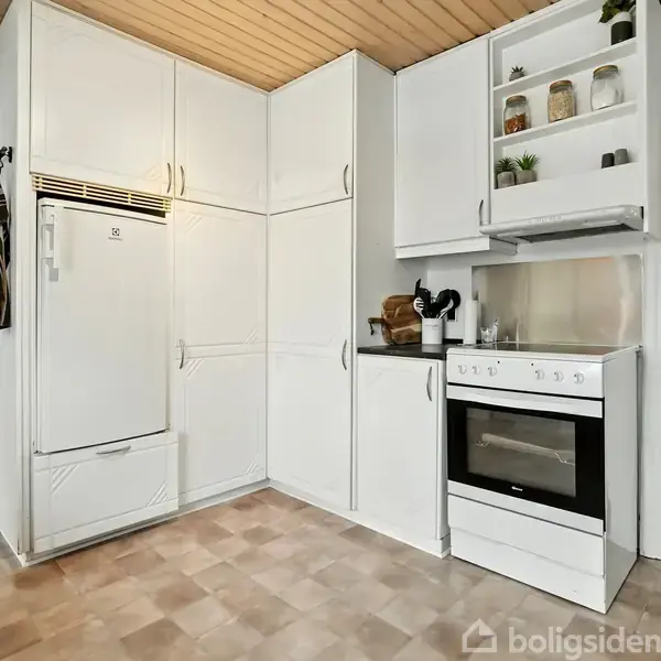 Køleskab står stille i et hvidt, rummeligt køkken med indbyggede skabe og en ovn. Trægulvet og det lyse loft skaber en lys atmosfære. Tilstødende rum ses gennem en døråbning.