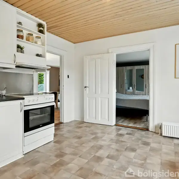 Et køkken med hvide skabe og en ovn, der står inaktiv. Et åbent dørhul fører til et tilstødende rum, og væggen har en indrammet billedkunst.