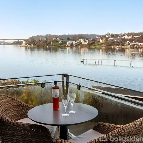 To fletstole ved et rundt bord med to glas og en flaske i en altan. Set over en skovklædt søbred med en bro i baggrunden.