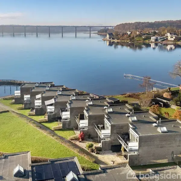 Boligblokke ligger langs en sø med spejlreflektioner. En bro krydser vandet i baggrunden, mens grønne områder omkranser husene. Træer og flere huse ses i landskabet.
