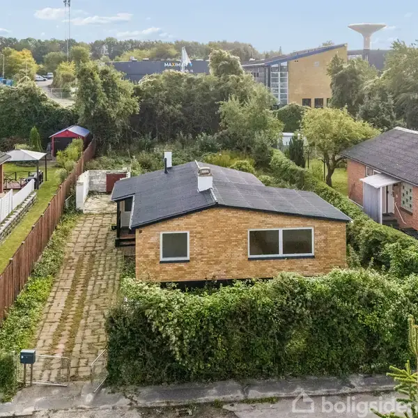 Huset er en enkelt etplans bygning med en flad sort tag, omgivet af hække i en villakvarter. Et flisebelagt indkørsel ligger til venstre. Omkringliggende bygninger er i samme stil.