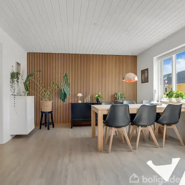 Et spisebord med fire sorte stole står ved et stort vindue. Grønne planter og en pendellampe skaber en hyggelig atmosfære i det lyse rum med trægulv og lamelvæg.