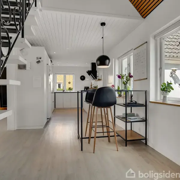 En moderne spisestue med høje stole ved et bord. Store vinduer slipper lys ind og en trappe leder til overetagen. Værelset er lyst med en skandinavisk indretning.