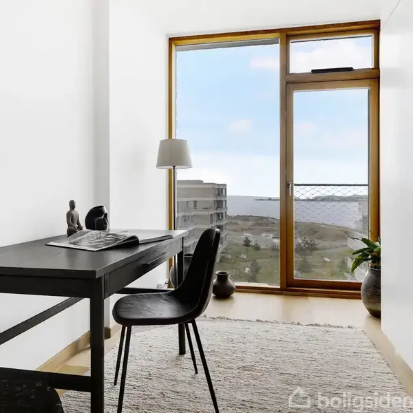 Et skrivebord står i et lyst rum med en stol foran. En lampe står på bordet, mens en stor vindue viser udsigt over vand og bygninger udenfor.