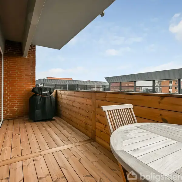 Træterrasse med rundbord og stol, omkranset af træhegn. En grill står i hjørnet op ad en murstensvæg. I baggrunden ses andre bygninger mod en blå himmel.