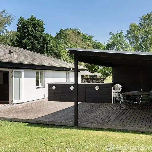 En overdækket terrasse med spisebord og stole står til højre, tilstødende et hvidt hus med åben skydedør til venstre, omgivet af frodig have og træer.