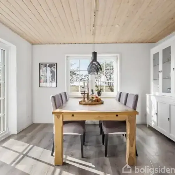 Træbord med seks stole står i et lyst spisestueområde. En sort pendellampe hænger over bordet. Rummet har store vinduer og trægulv. En hvid skænk står til højre.