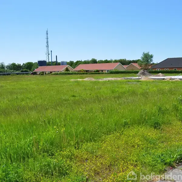 Grønt græsareal strækker sig foran et boligområde med huse i horisonten under en klar blå himmel. En høj telemast står blandt husene i baggrunden.