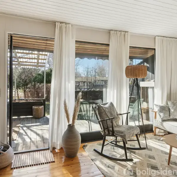 En stue med hvide gardiner hængende ved store glasdøre, der åbner ud til en terrasse. Et bord og stol står på et lyst trægulv. Indretningen er lys og moderne.