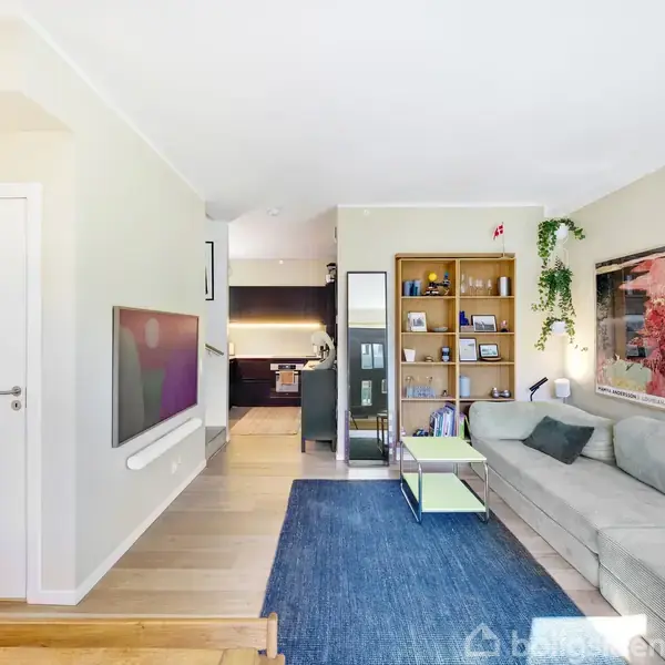 En moderne stue med en grå sofa langs væggen, udsmykket med to plakater. Reoler fyldt med bøger og dekorativt grønt. Et stort TV hænger på den lyse væg; køkken i baggrunden.