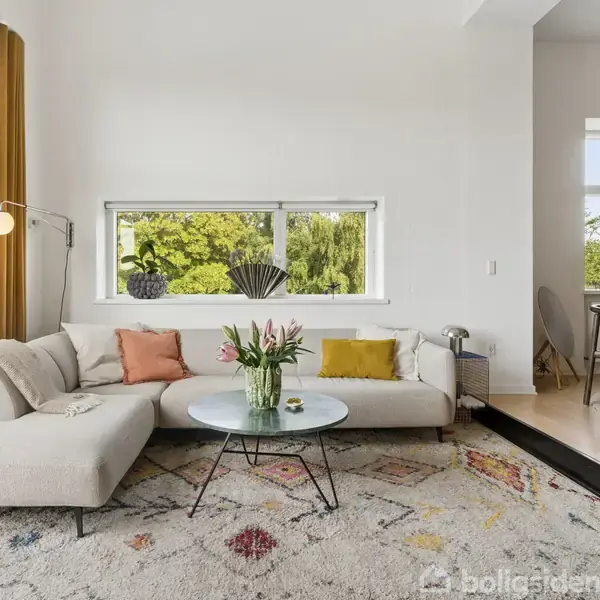 En beige sofa med farvede puder står i en lys stue ved siden af et rundt sofabord med blomster. Store vinduer og en dør åbner ud til en terrasse med grøn udsigt.