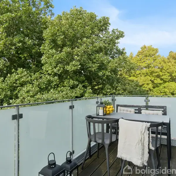 En balkon med bord og stole er placeret mod en frodig, grøn skovbaggrund. Der er lanterner og en skål med citroner på bordet, omgivet af en glasafskærmning.