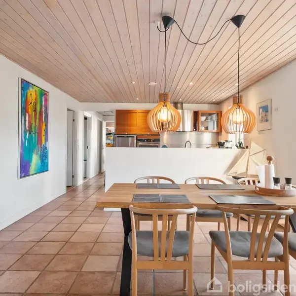 Spisebord med stole står i et åbent, lyst køkken-alrum. Loftet har træpaneler, gulvene er flisebelagte, og der hænger kunst på væggen. Over bordet hænger moderne lamper.