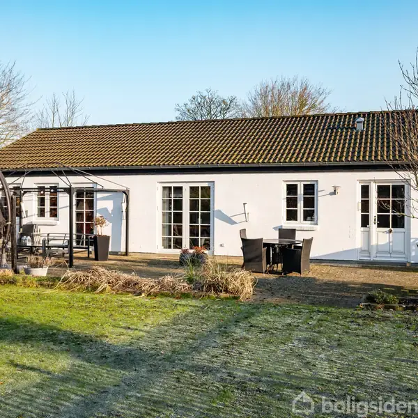 Hus med hvid facade og teglstenstag, står i en solrig have. Der er en terrasse med borde og stole, omgivet af græsplæne og træer i baggrunden.