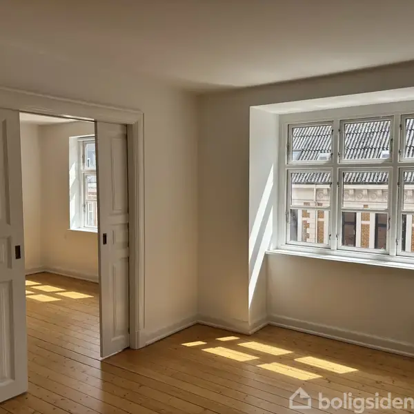 Et lyst rum med åbne skydedøre afslører trægulve og et vindue med udsigt over en gade kantet af bygninger. Sollys strømmer ind i rummet.