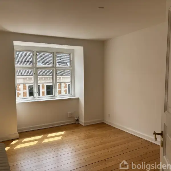 Et lyst værelse med trægulv og hvide vægge. Et vindue med udsigt til en murstensbygning lukker sollys ind, og en uåbnet dør står til højre i rummet.