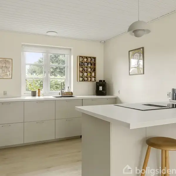 Et moderne køkken med et hvidt køkkenbord og to træstole. Køkkenskabe og apparater er placeret i baggrunden. Væggene er dekoreret med kunst og en krydderihylde ved vinduet.