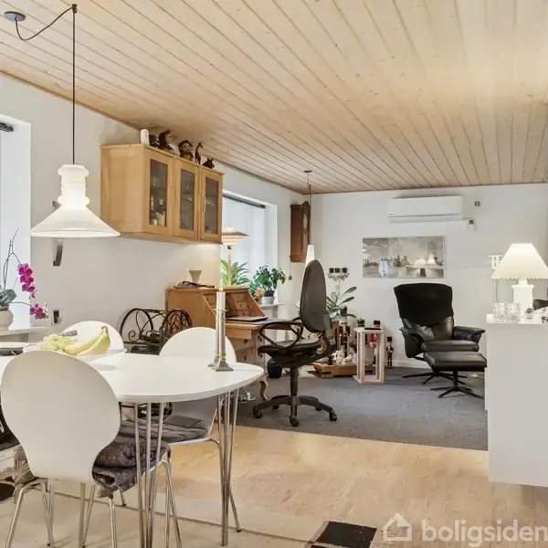 Et lyst og indbydende spisestueområde med hvide møbler og spisebord. Til højre ses en sort lænestol og skrivebordstol i en stue med lyse vægge samt træloft. Indendørsplanter pynter rummet.