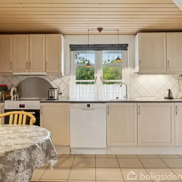 Et lyst køkken med træloft og lyse træskabe. En opvaskemaskine er centreret under et vindue. Et rundt bord med dug står til venstre. Kvadratiske fliser på gulvet.