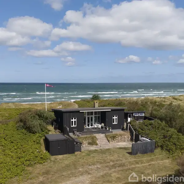 Et sort sommerhus står stille omgivet af klitter og frodig vegetation nær kysten med havudsigt. En dansk flagstang er placeret ved siden af huset under en klar himmel.