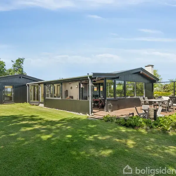 En enkelthistorisk hus med terrasse står midt i en frodig have. Der er havemøbler på terrassen, omkranset af buskads, mens grønne træer omgiver ejendommen under en klar blå himmel.