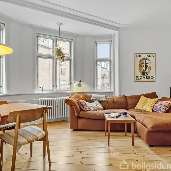 Brun hjørnesofa står i stue med lyse trægulve og store vinduer. Et spisebord med stole står til venstre. Plakat på væggen siger "LOUISIANA PICASSO". Bøger og planter pynter rummet.