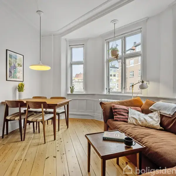 En stue med en brun sofa placeret ved træbord med en kop. Der er et spisebord med fire stole, maleri på væggen og store vinduer, der giver naturligt lys.