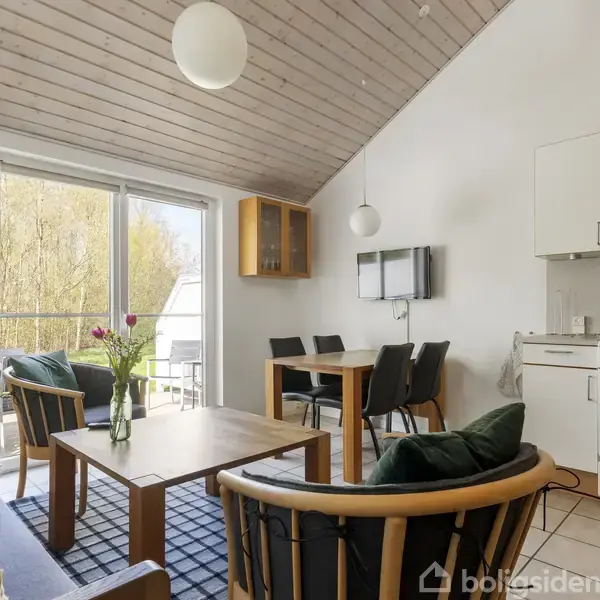 En stue med sofa, lænestole og sofabord, arrangeret på et rektangulært tæppe. Et spisebord står for enden. Et åbent køkken er til højre, med en ovn. Store vinduer giver udsigt til træer udenfor.