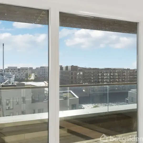 Store glaspartier viser en udsigt over en moderne bydel med bygninger og en skorsten. Solskin oplyser altanen udenfor.