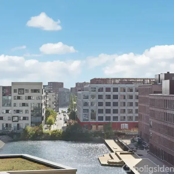 Flod omgivet af moderne bygninger, hvor floden forløber gennem et byområde med blå himmel og spredte skyer i baggrunden.