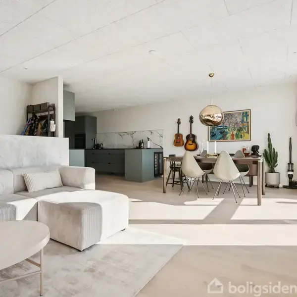 En moderne stue med lys sofa og rundt bord i forgrunden. I baggrunden ses et spisebord med stole, guitarer hængende på væggen, planter og kunstmaleri. Ingen synlig tekst.