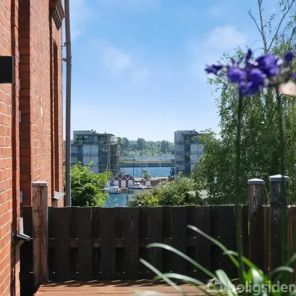 Murstensbygning står stille ved siden af en træhegn, mens en lilla blomst svajer let i forgrunden; baggrunden viser moderne bygninger ved vandet under en klar blå himmel.