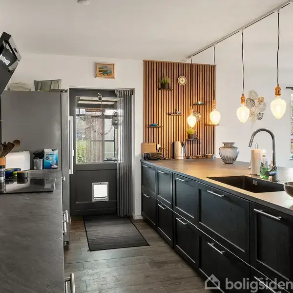 Moderne køkken med sorte skabe på begge sider og en række dekorative lamper hængende over bordpladen. En dør og et vindue i baggrunden giver udsigt til haven.