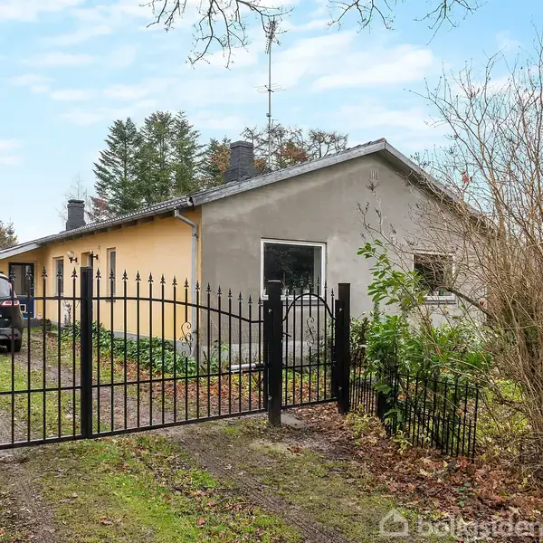 Et gult hus med gråt tag står bag en sort metalport. En bil holder i indkørslen. Omgivet af træer og græsplæne under en skyet blå himmel.