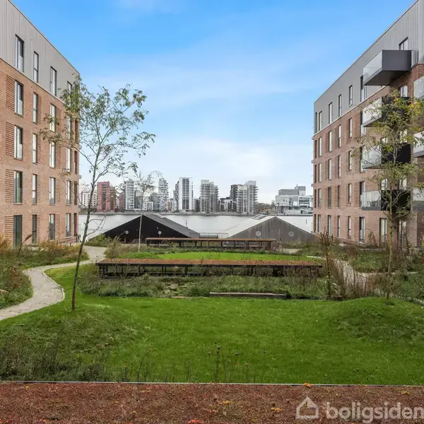 To moderne murstensbygninger med altaner står overfor hinanden, omkranser en grøn gårdhave med stier og træer. I baggrunden ses en bysilhuet mod en blå himmel.