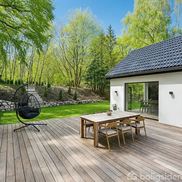 Terrasse med træbord og stole i en have med grønne træer og græsplæne. Et moderne hus med store glasdøre ses i baggrunden. En hængestol står på terrassen.