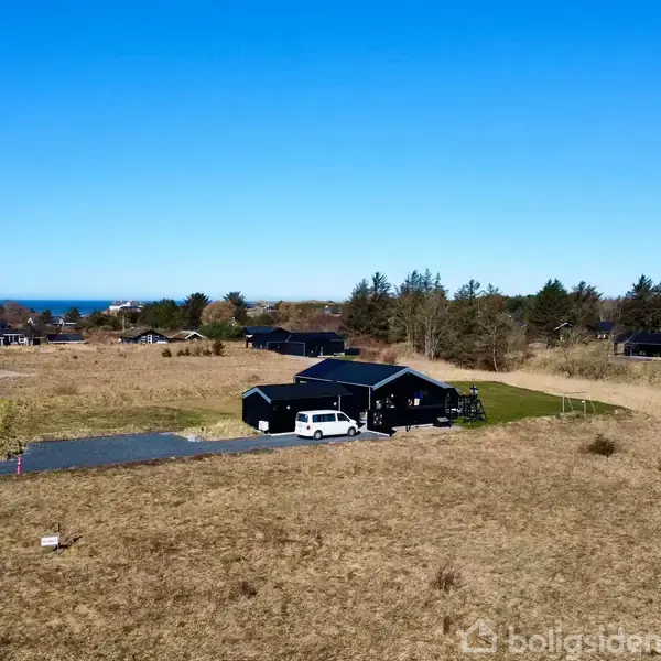 Sort hus omgivet af vildt, tørt græs med en parkeret hvid bil foran. Beliggende i et landligt område tæt på skov og kystlinje i baggrunden.