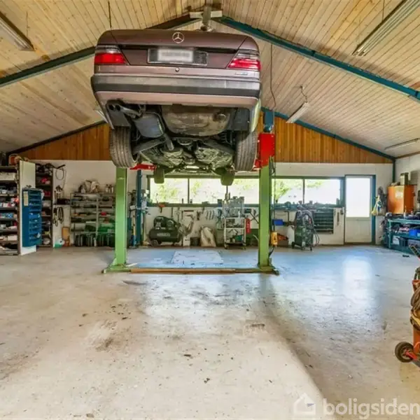 Bil hævet på en hydraulisk lift i en fyldt autoværksted, omringet af værktøjer og udstyr i hylder og skabe; stort vindue i baggrunden lader naturligt lys ind.