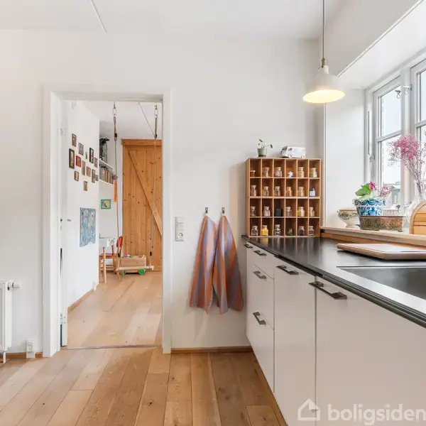 Køkkenbord med vask har viskestykker hængende ved siden af og en vinreol ovenpå; rummet er lyst med et trægulv. I baggrunden ses en åbning til en pyntet gang.